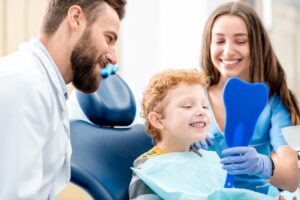Dentist working with child smiling into mirror while parent sits beside him.